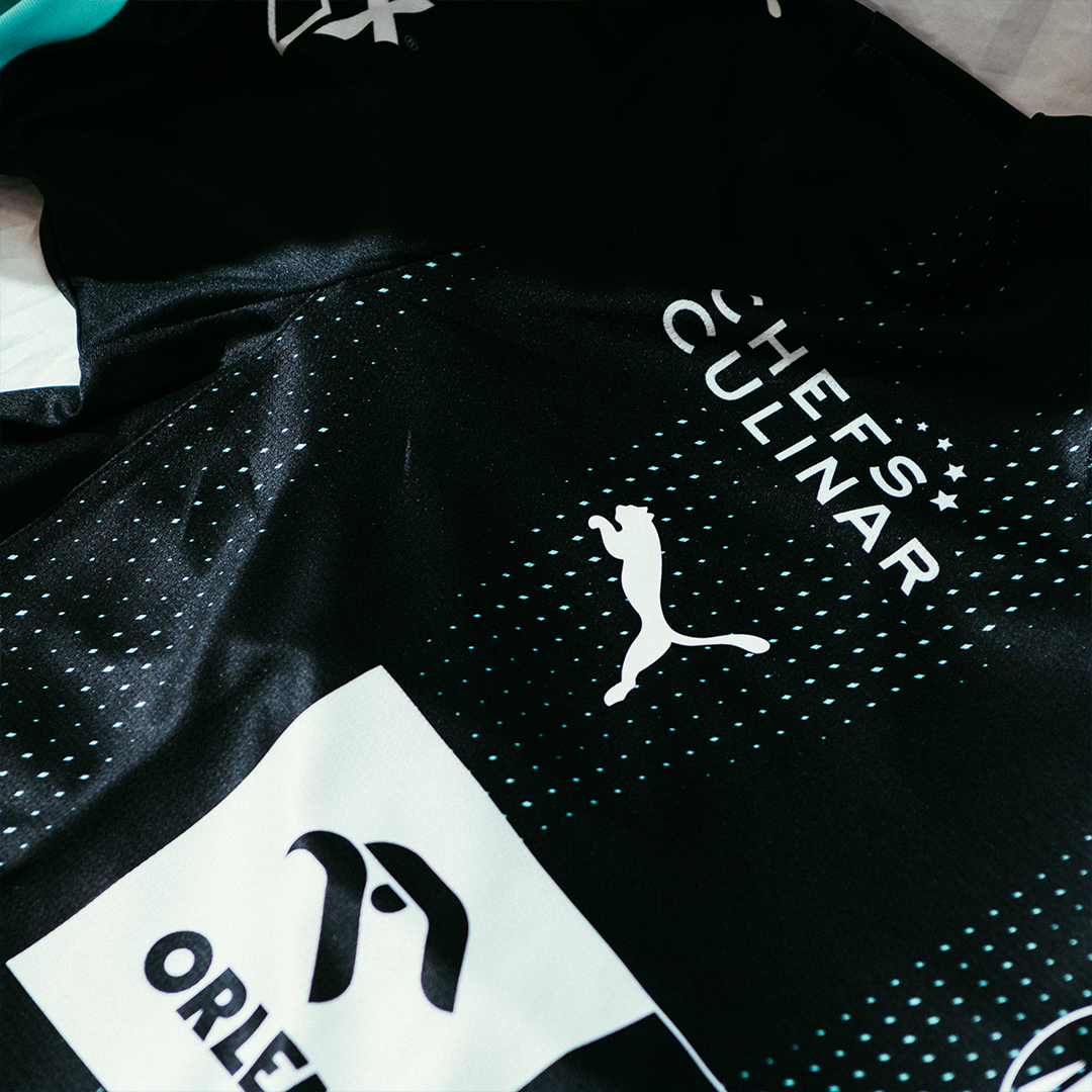Eine Nahaufnahme des THW Kiel 24-25 Auswärtstrikots in PUMA-Schwarz zeigt das Puma-Logo, den Schriftzug CHEFS CULINAR, ein teilweise sichtbares Orlen-Logo mit einem Vogelmotiv sowie weiße und blaugrüne Akzente auf dem Stoff.