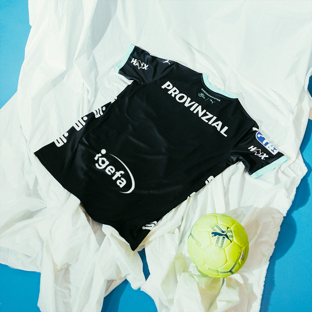 Ein THW Kiel 24-25 Auswärtstrikot PUMA Schwarz mit weißen Sponsorenlogos liegt auf einem weißen Blatt neben einem gelb-grünen Handball auf einer blauen Fläche.