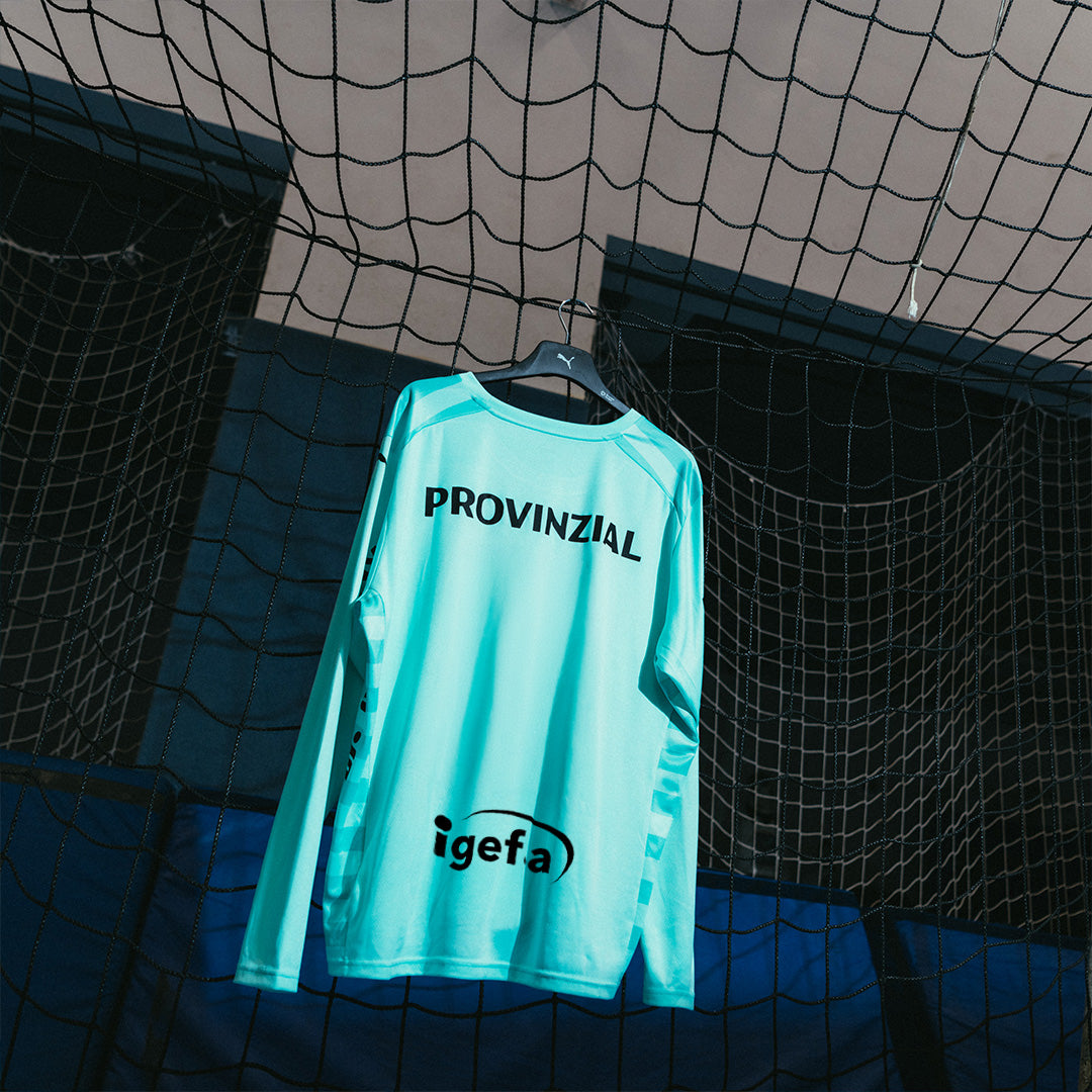 Das THW Kiel 24-25 GK Jersey Home aus recyceltem Polyester in Teal mit "PROVINZIAL" und "igefa" Logos hängt am Netz im Indoor-Sportbereich, umgeben von Netzstrukturen und blauen Hallenwänden.