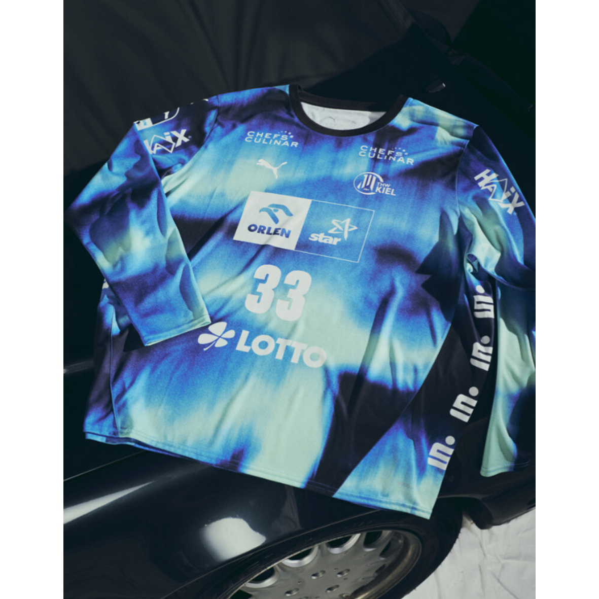 Ein THW Kiel 25-26 GK Trikot Home by PUMA in navy, mit Sponsorenlogos und der Nummer 33, liegt auf der Motorhaube eines schwarzen Autos, auf dem ein Teil eines silbernen Rades zu sehen ist.