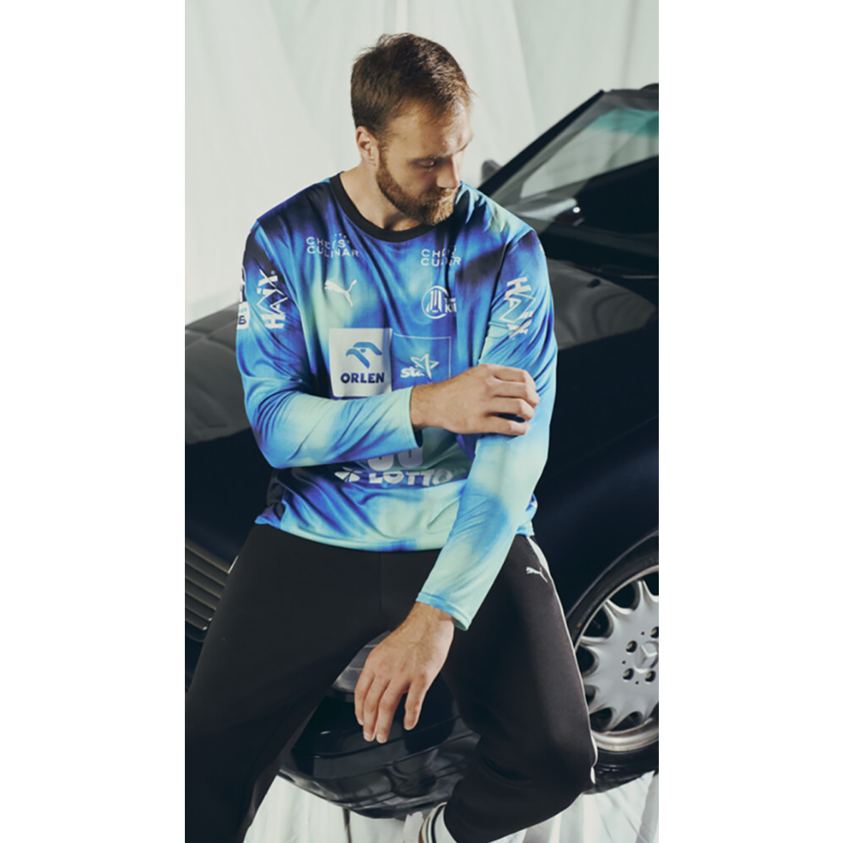 Ein bärtiger Mann sitzt auf einer schwarzen Autohaube, schaut nach unten und rückt den Ärmel seines blauen THW Kiel 25-26 GK Trikots Home PUMA Navy mit Sponsorenlogos zurecht. Er trägt eine schwarze Hose mit weißen Streifen.