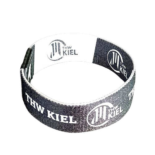 Das Elastische Armband schwarz/weiß ist ein schwarz-weißes RPET-Armband mit dem Schriftzug "THW KIEL" und einem kreisförmigen Logo, das wiederholt auf schwarzem Hintergrund aufgedruckt ist.